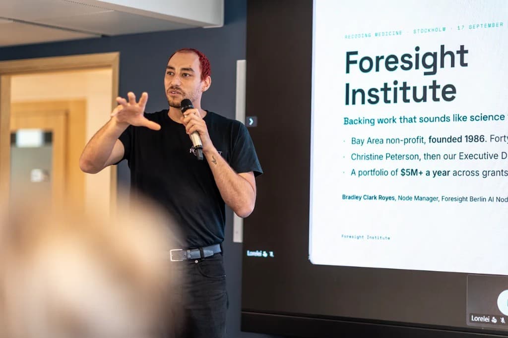 Bradley Clark Royes speaking at Recoding Medicine in Stockholm, microphone in hand, Foresight Institute slide behind him