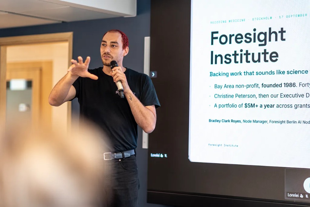 Bradley Clark Royes speaking at Recoding Medicine in Stockholm, microphone in hand, Foresight Institute slide behind him